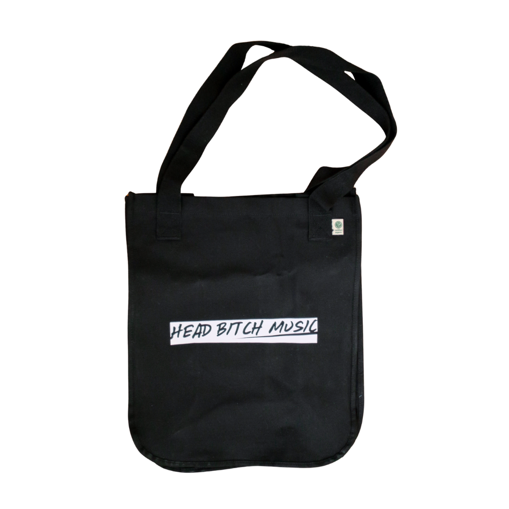 A black canvas tote bag with a single shoulder strap, featuring a white rectangular printed logo on the front with the text 'HEAD BITCH MUSIC' in black handwritten-style lettering and a white printed text on the back with the text 'BITCHES GET PAID'