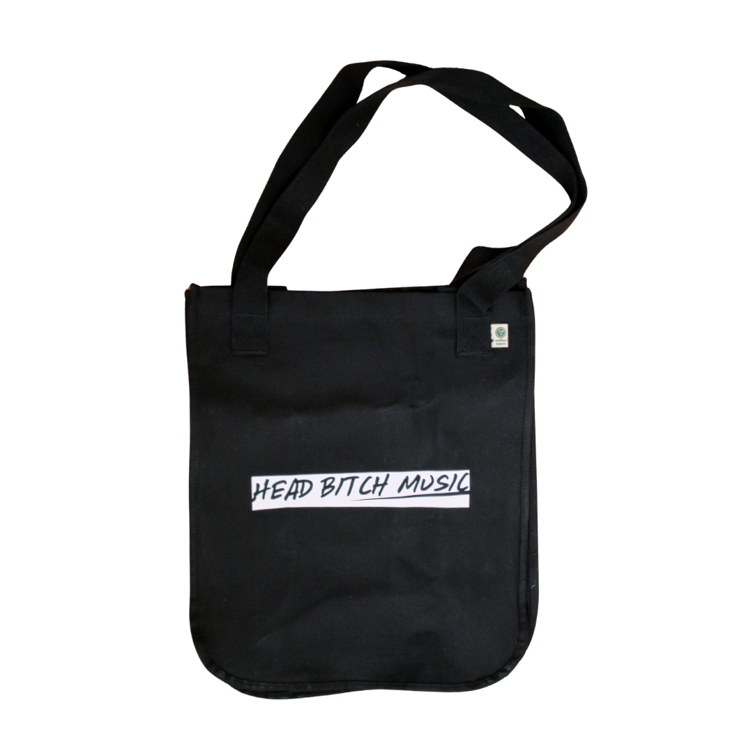 A black canvas tote bag with a single shoulder strap, featuring a white rectangular printed logo on the front with the text 'HEAD BITCH MUSIC' in black handwritten-style lettering and a white printed text on the back with the text 'BITCHES GET PAID'